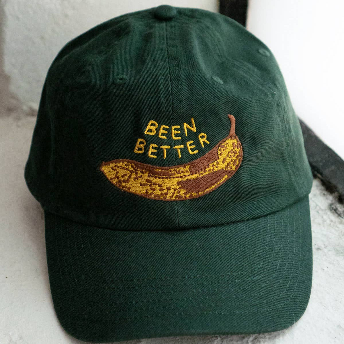 Green cap with a banana graphic and 'BEEN BETTER' text on a white surface.
