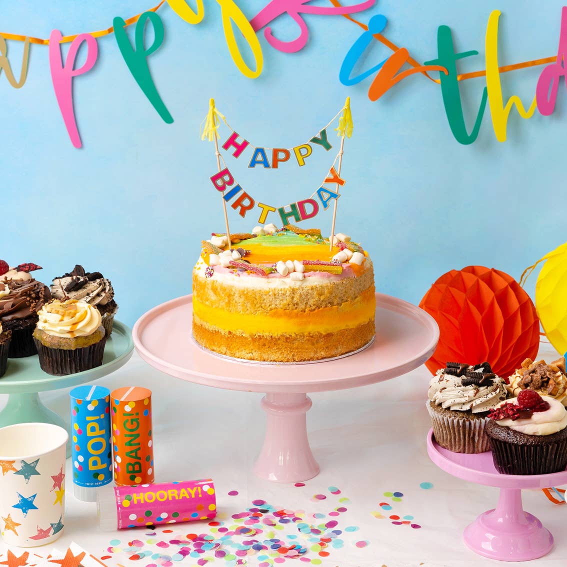 Birthday cake with 'Happy Birthday' candles, cupcakes, and confetti on a table with a colorful 'Happy Birthday' banner in the background.