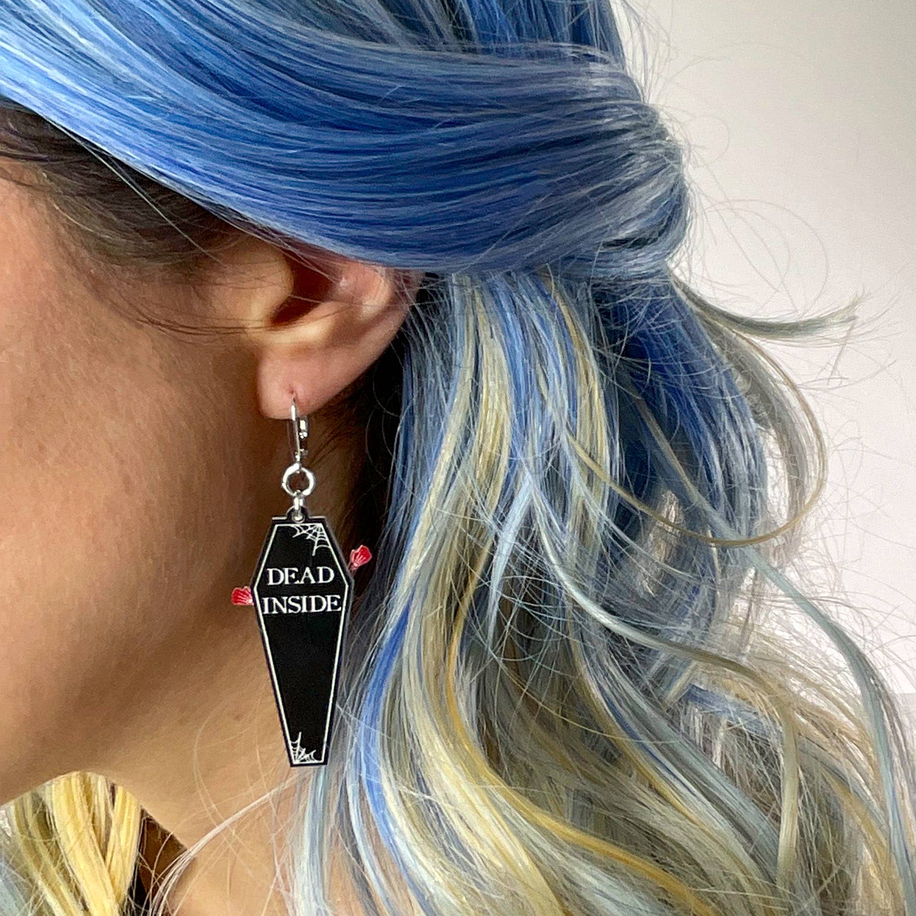 Person with blue and blonde hair wearing a coffin-shaped earring with 'Dead Inside' text.