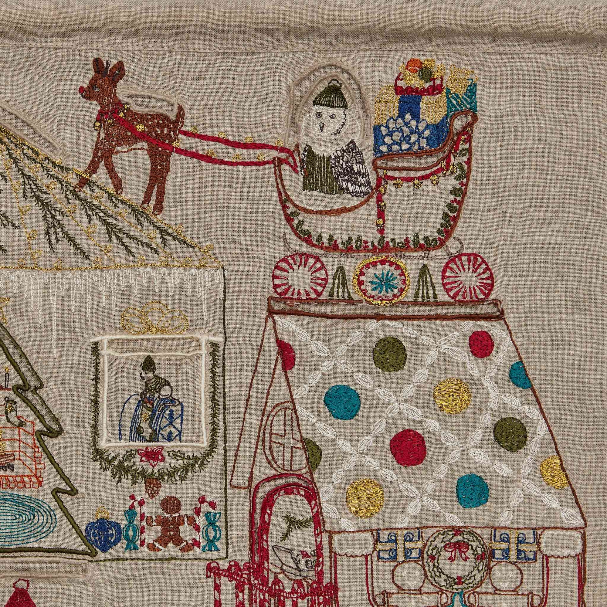 Embroidered scene with a reindeer, sleigh, and gingerbread house on textured fabric.