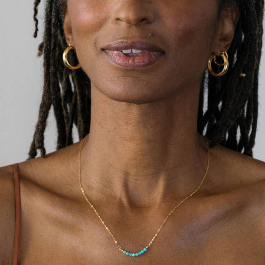 Woman wearing gold hoop earrings and a turquoise necklace against a neutral background