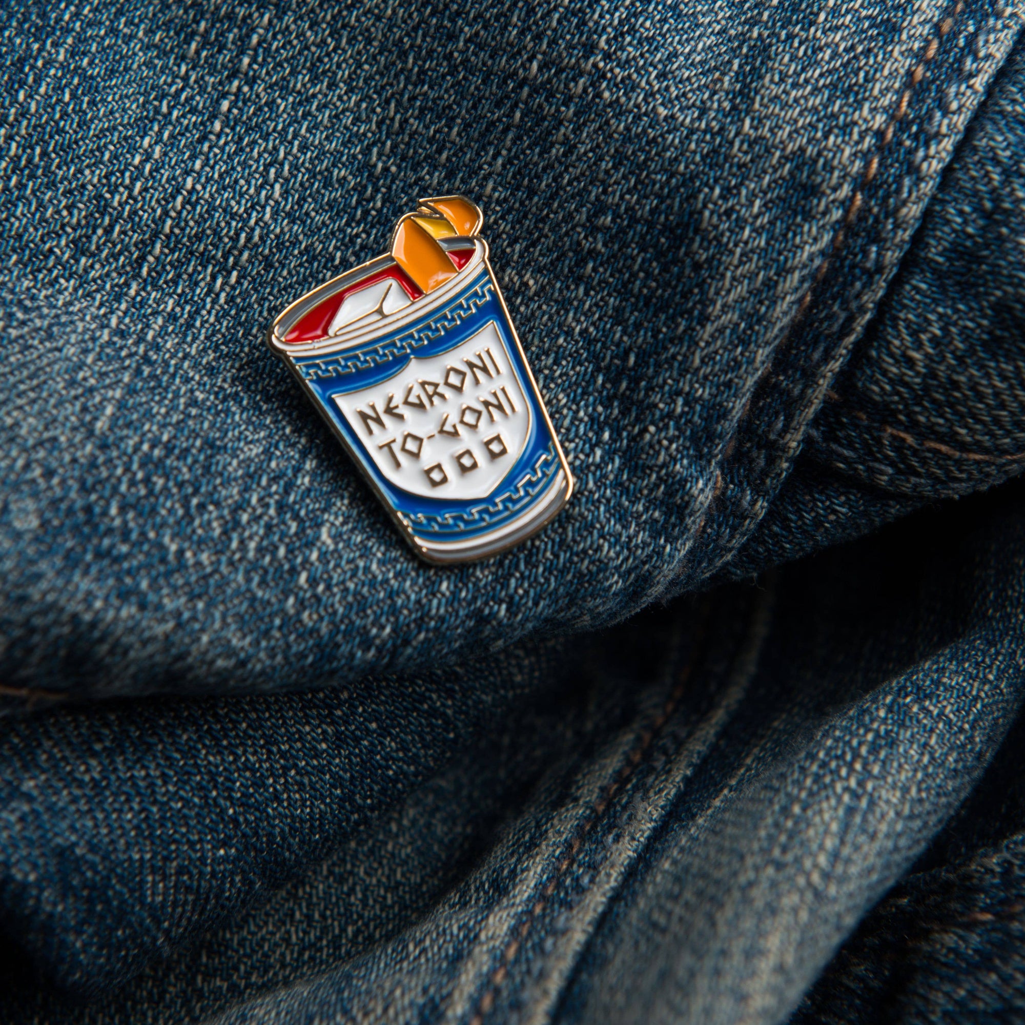 Enamel pin of a can labeled 'Negroni to Go!' on denim fabric