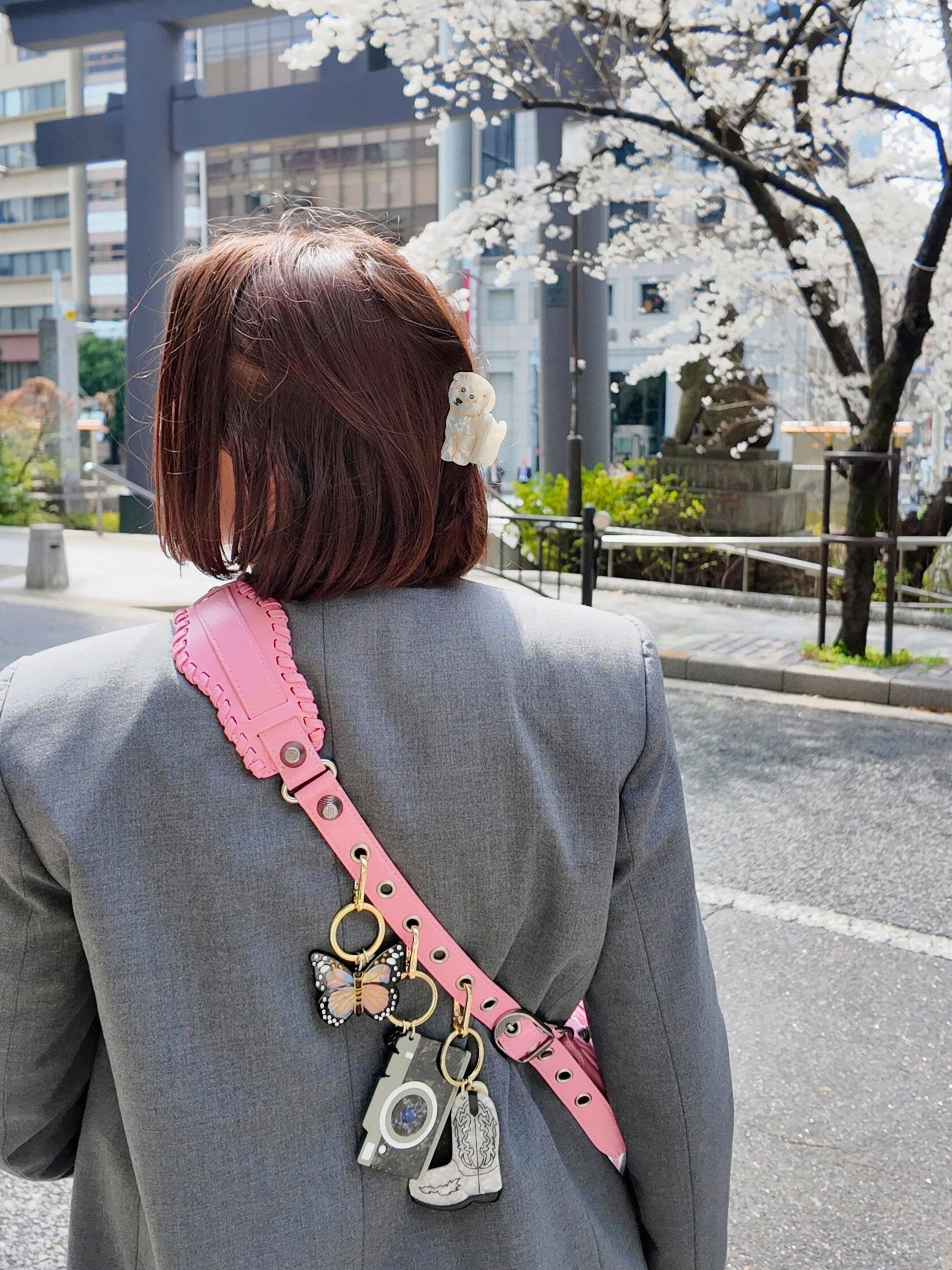 Person wearing a gray blazer with a pink strap and charms, standing in an urban setting with cherry blossoms.