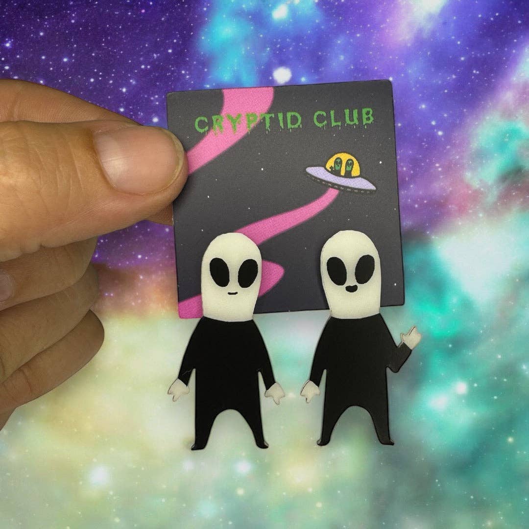 Alien-shaped earrings held in front of a 'Cryptid Club' card with a colorful background
