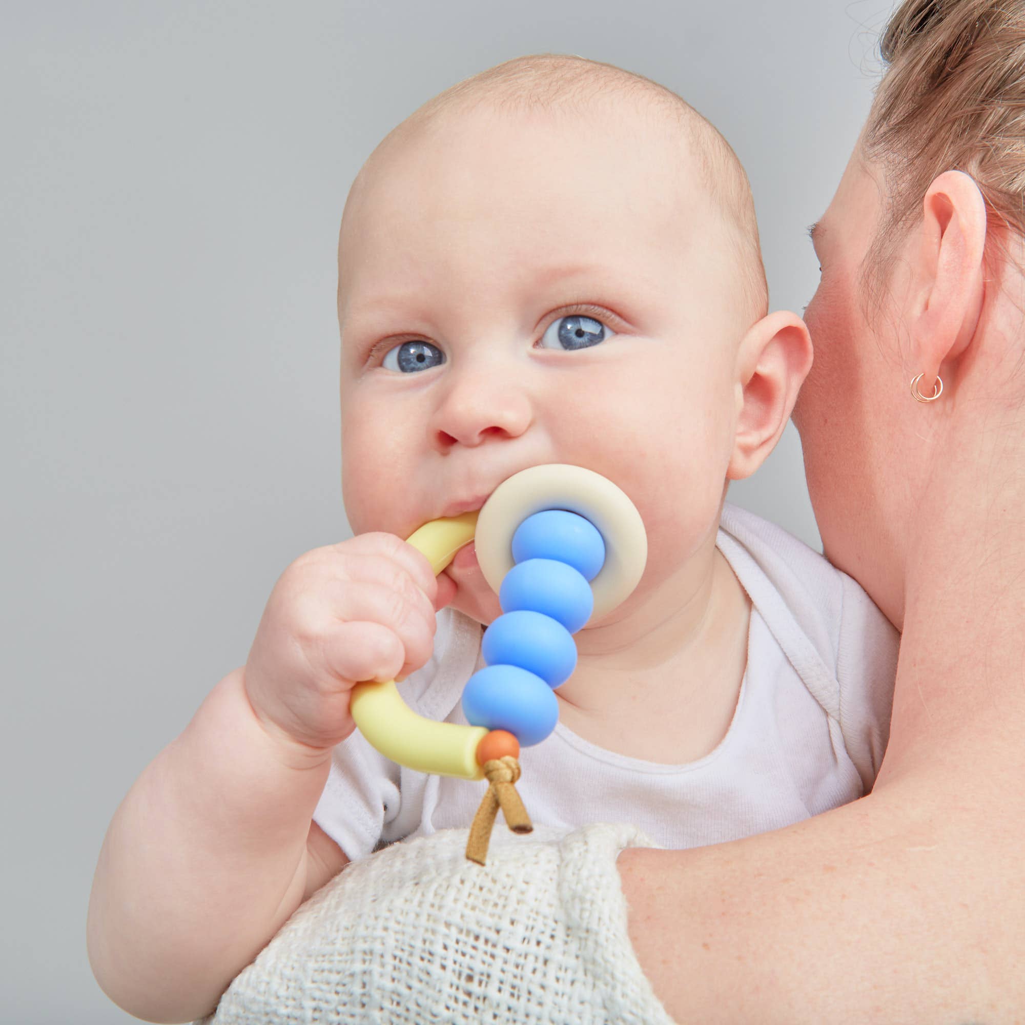 Baby holding a colorful teething ring with a person holding the baby against a gray background