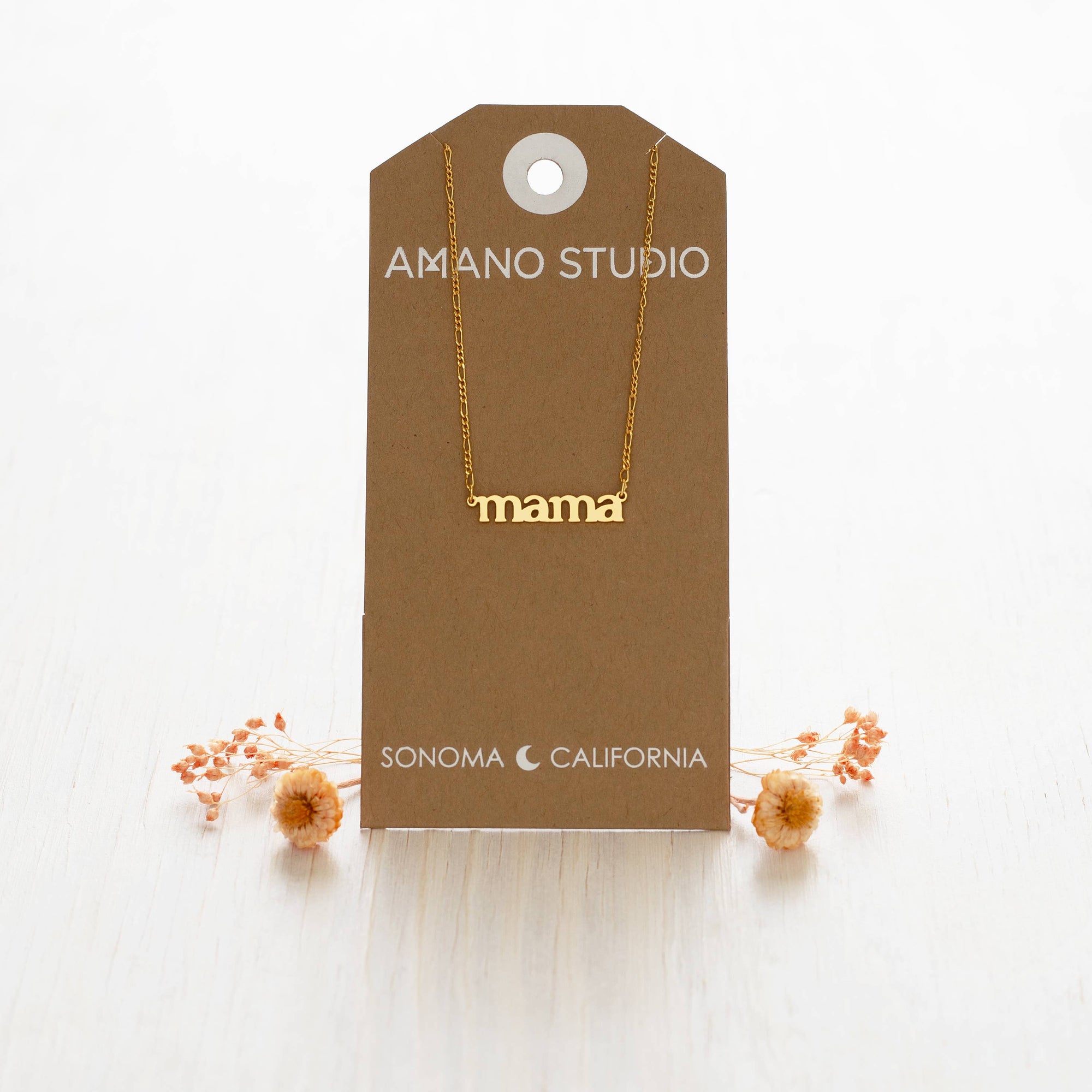 Gold 'mama' necklace on a brown AMANO STUDIO card with dried flowers on a white background