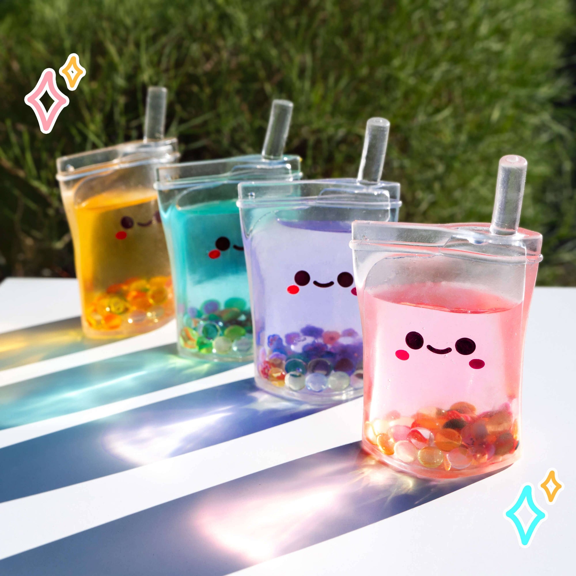 Four transparent cups with colorful contents and smiling faces on a reflective surface with greenery in the background.