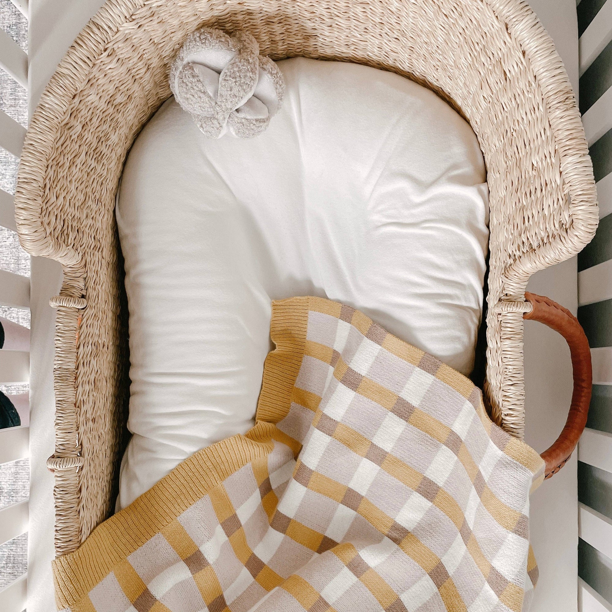 Baby crib with white bedding, checkered blanket, and baby shoes on a striped background