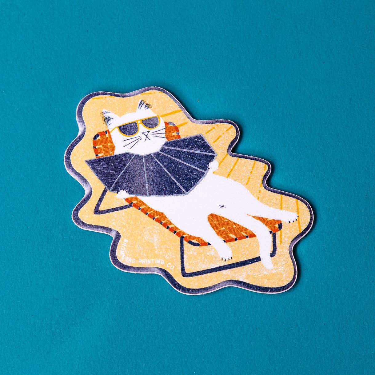 A mirror sticker with an illustration of a cat wearing sunglasses, lounging in a chair, with a reflector.