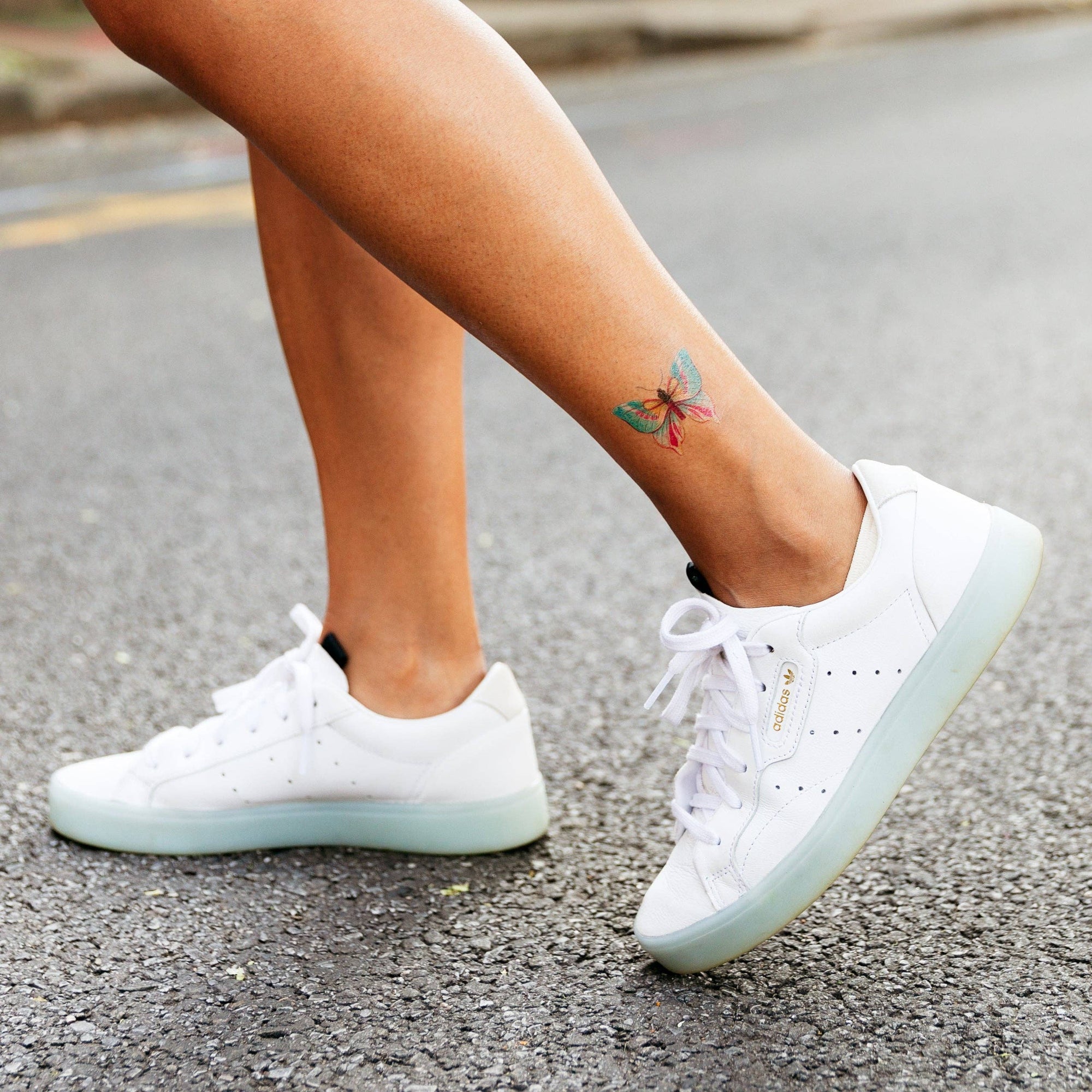 White sneakers worn on a person's feet with a colorful tattoo on the ankle.