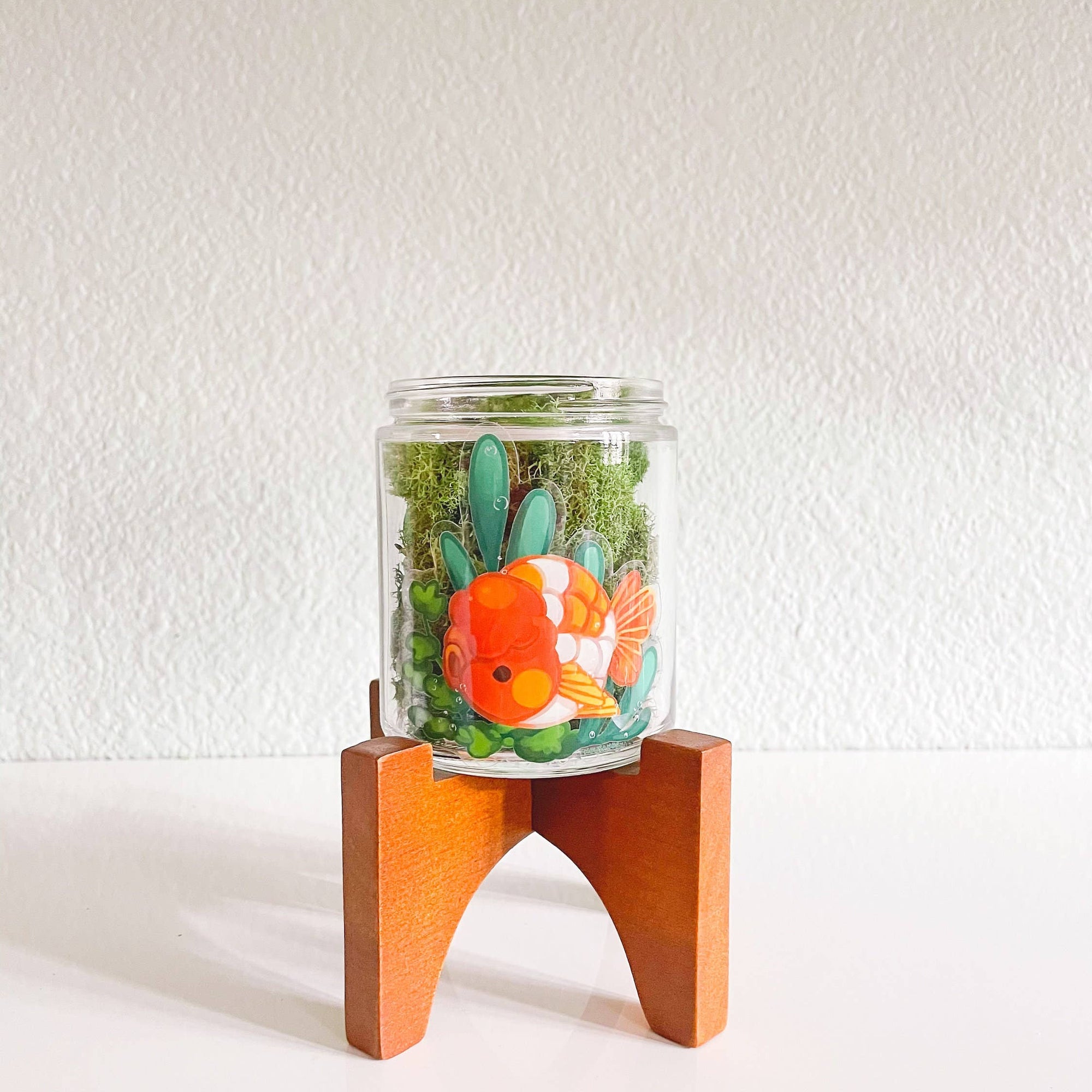Glass jar with a decorative fish and plants on a wooden stand against a white background