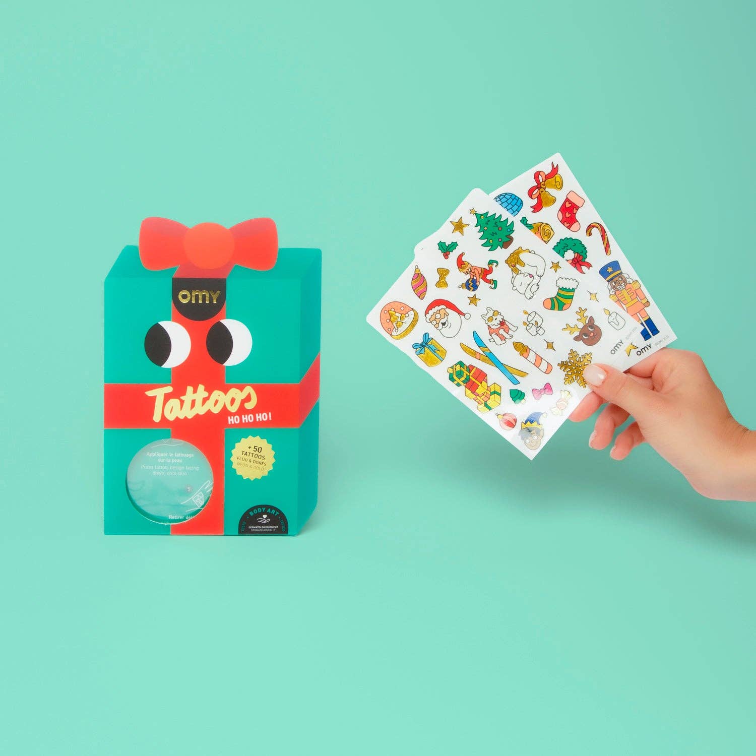 Hand holding a sheet of temporary tattoos next to its packaging on a teal background