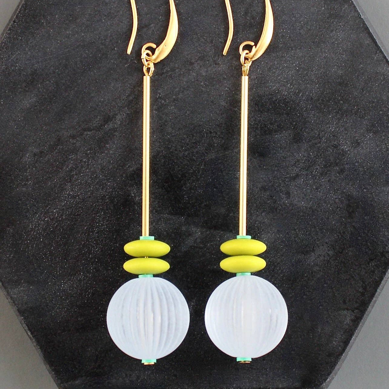 Pair of earrings with gold hooks, white spherical beads, and yellow accents on a black hexagonal stand.