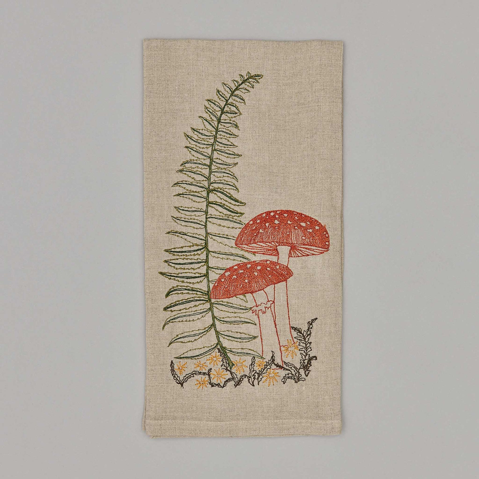 A beige cotton tea towel with a green and red embroidered design featuring a lady fern and an agaric mushroom.