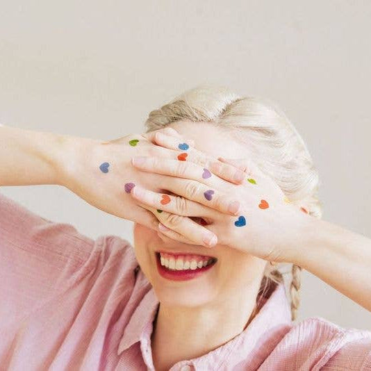 Person with colorful heart tattoos on hands covering face, wearing a pink shirt.