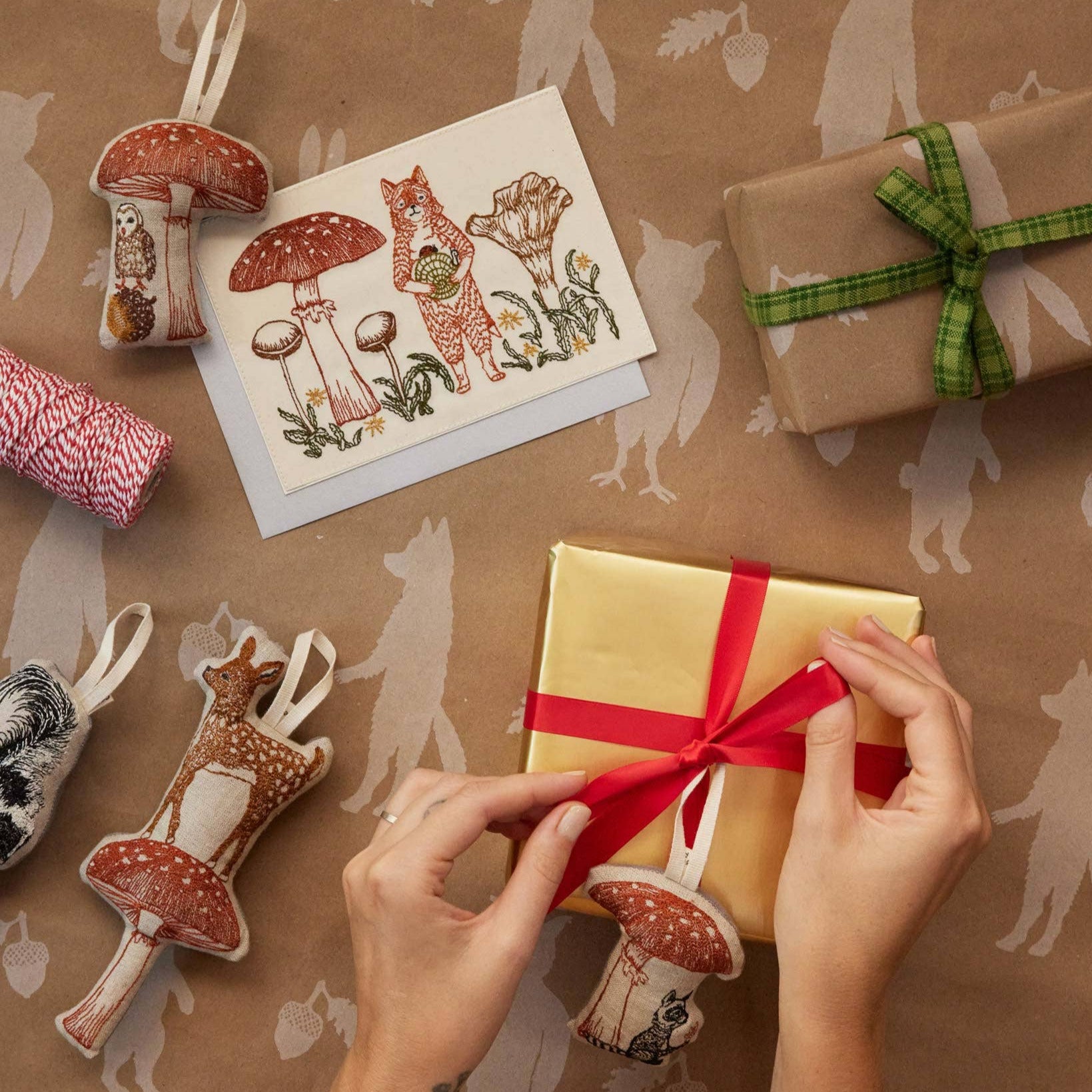 Person wrapping a gift with a red ribbon on brown paper with forest-themed decorations.