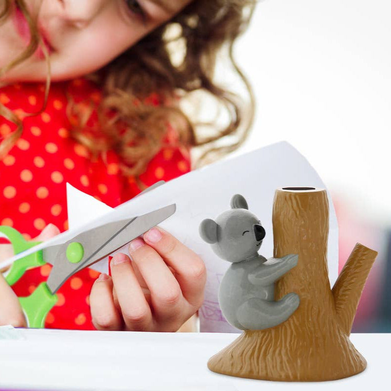 Child cutting paper with scissors next to a small koala figurine on a tree branch