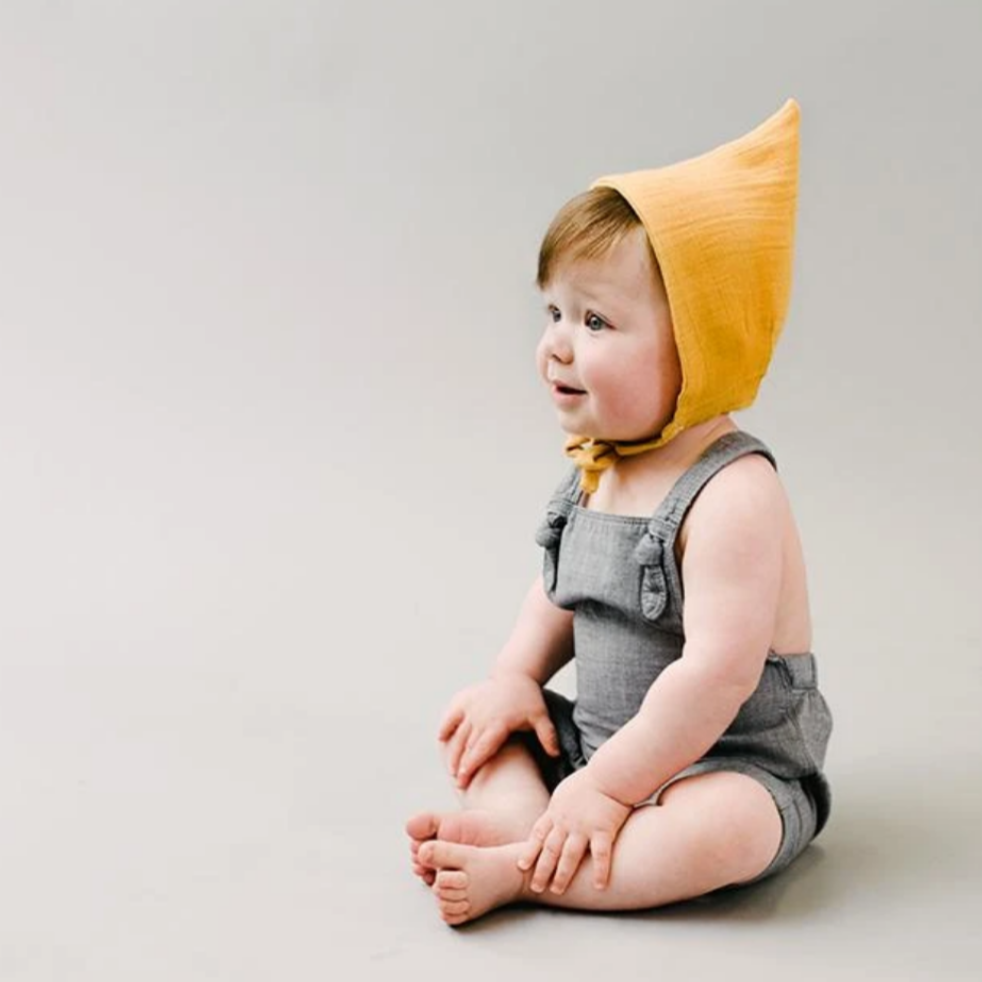 baby in grey overalls wearing mustard pixie bonnete