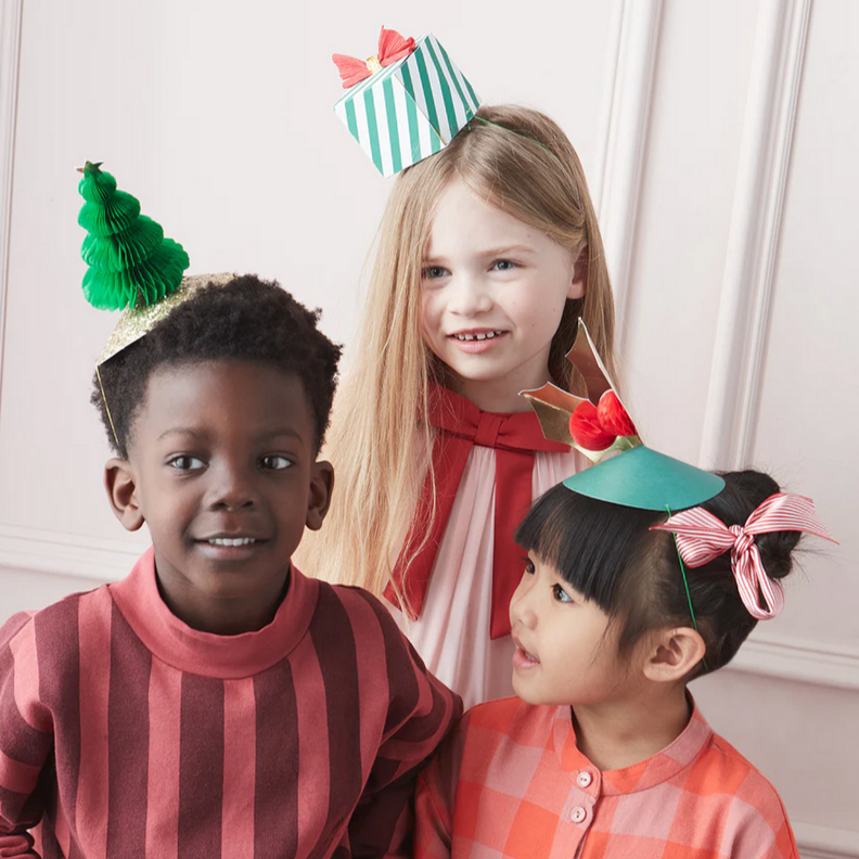 Three children wearing a variety of festive party hats with Christmas tree, holly, and gift designs in red, green, and gold colors.