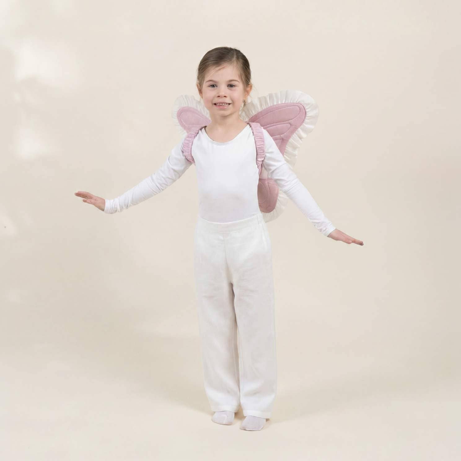 A young girl stands on a plain beige studio background wearing a white leotard and white pants, with pink fairy wings attached behind her shoulders.
