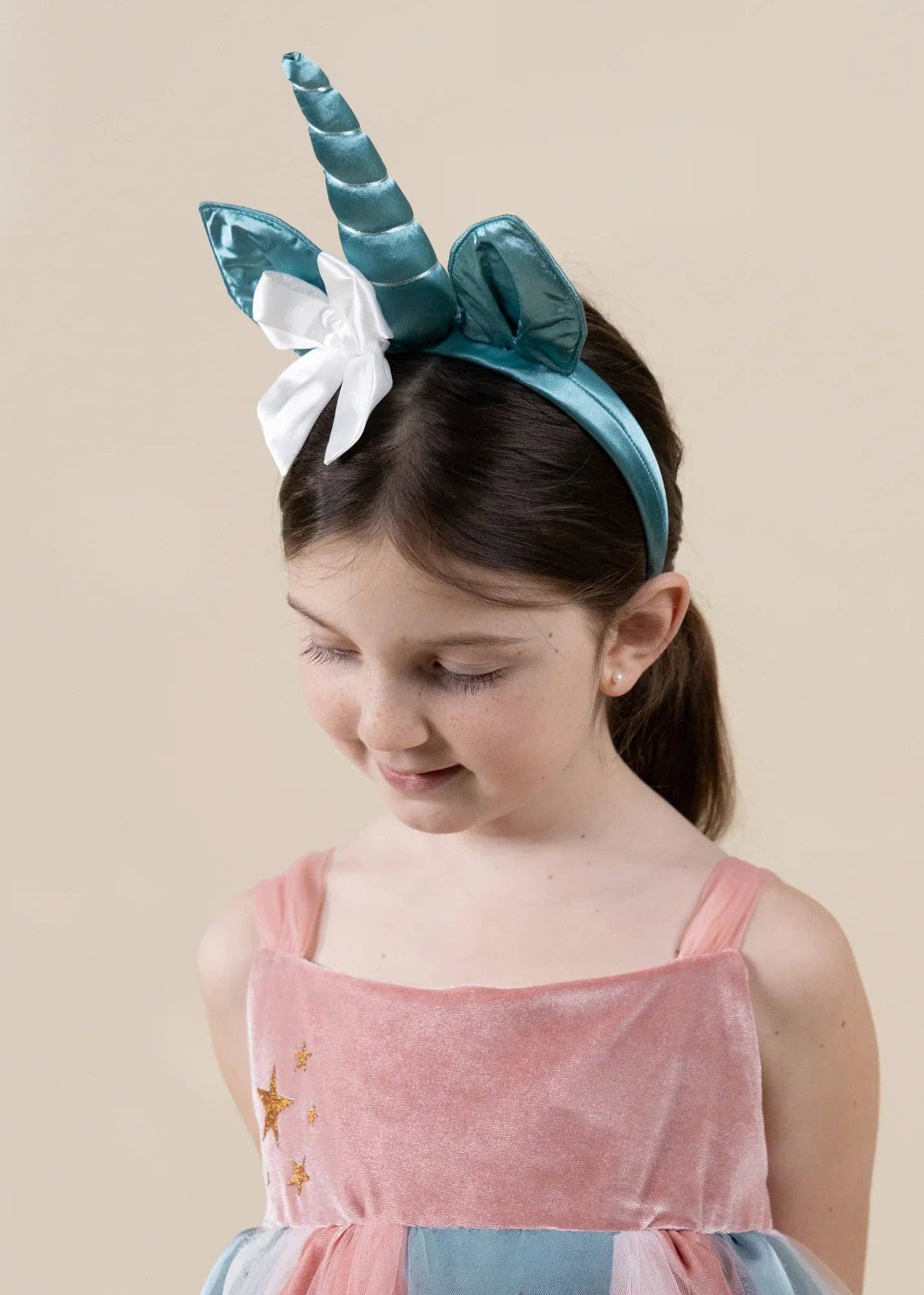 A young girl wearing a shiny teal unicorn headband with a white bow, paired with a pink dress, against a solid beige background.