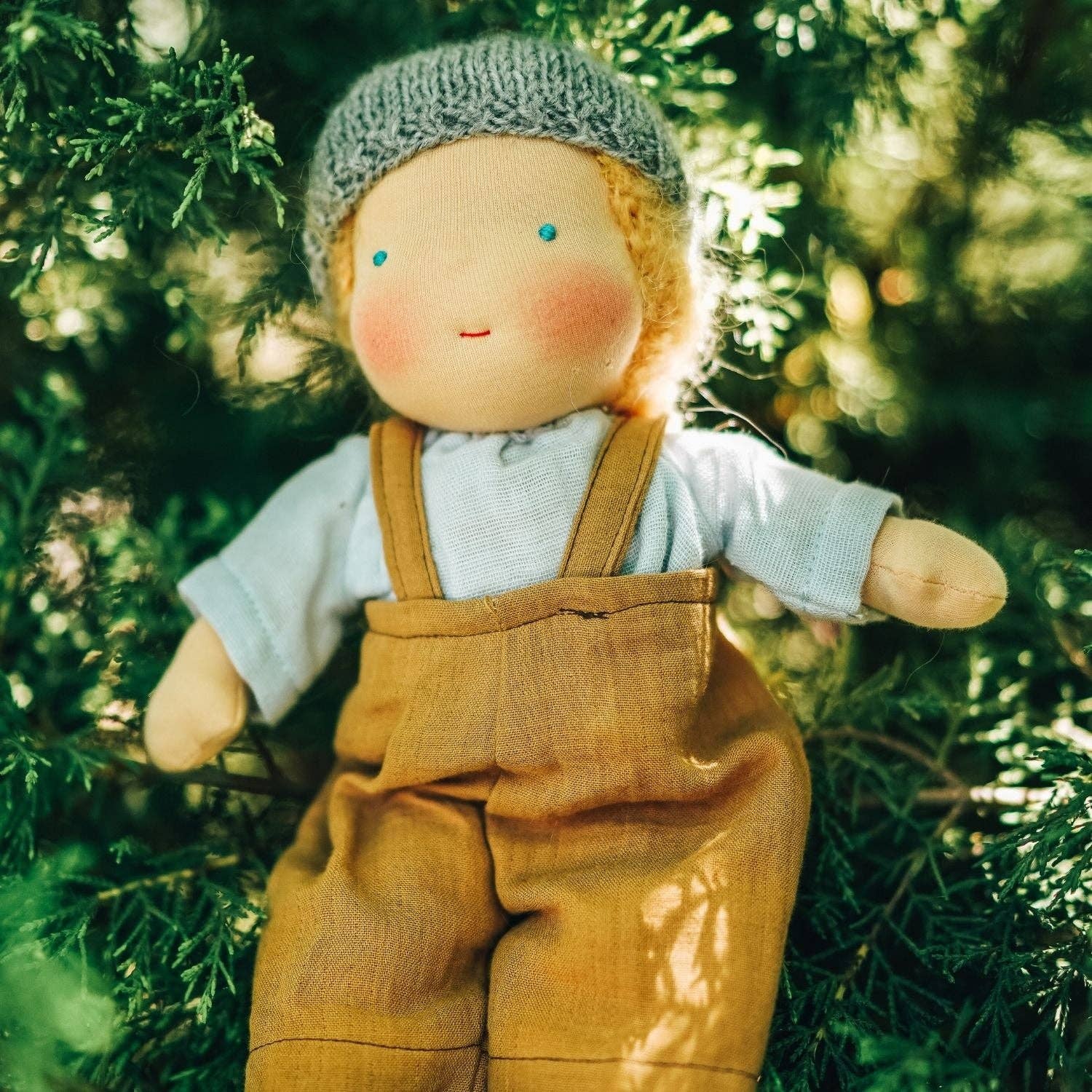 Doll wearing a knitted hat and overalls standing among green foliage