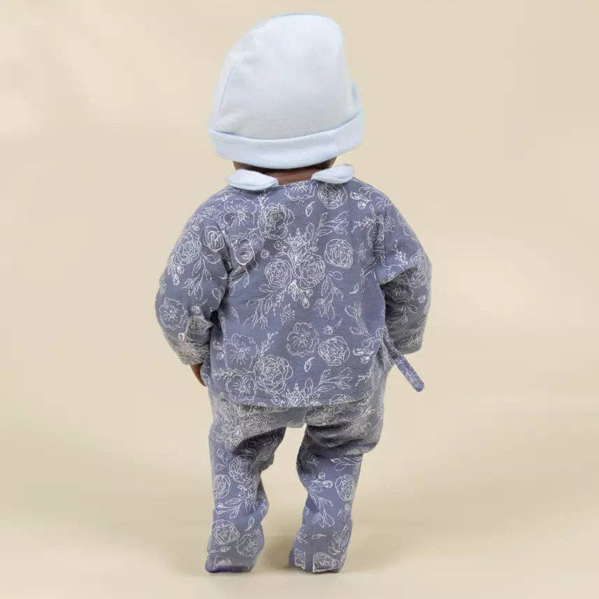 A baby viewed from behind wearing a light blue knit bonnet and a dusty blue floral patterned footed romper, set against a solid beige studio background.