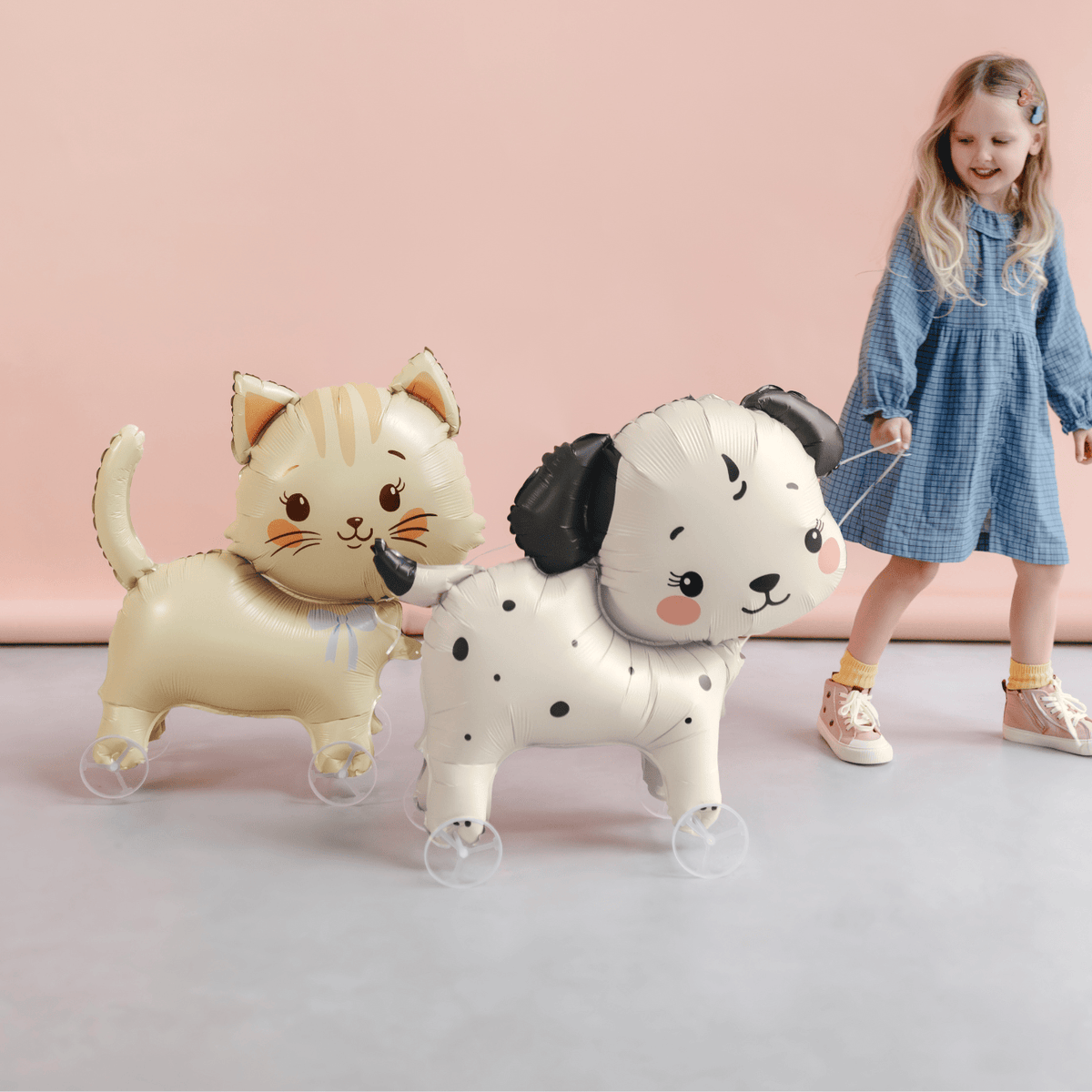 Child standing next to two inflatable animal balloons shaped like a cat and a dog on a pink background.