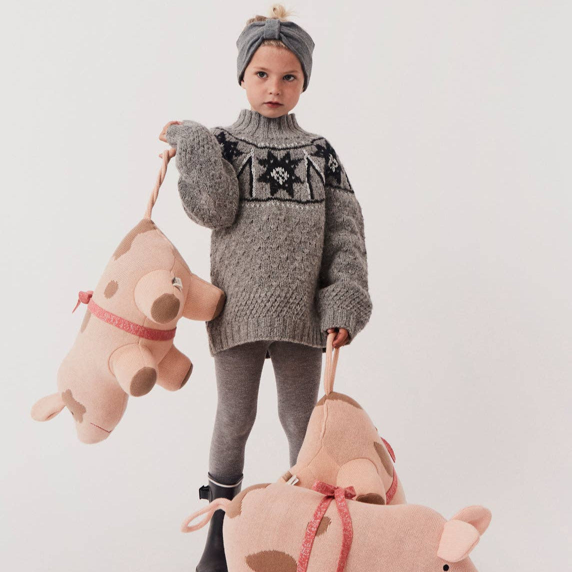 Child wearing a gray knitted sweater with star pattern, holding two pink stuffed pigs against a white background