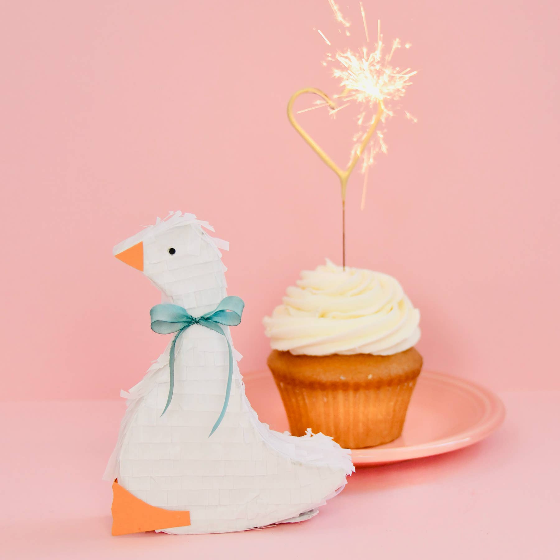 Stuffed goose with a bow next to a cupcake with a sparkler on a pink background