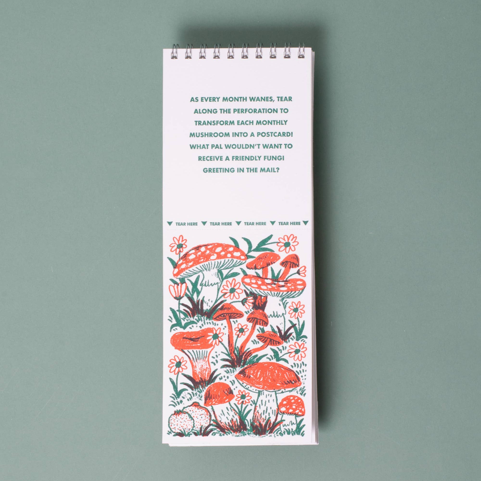 A tall, narrow spiral-bound notebook or desk calendar with a white cover, featuring a printed mushroom illustration in coral red, green, and black, and a short quote at the top, photographed on a muted sage green background.