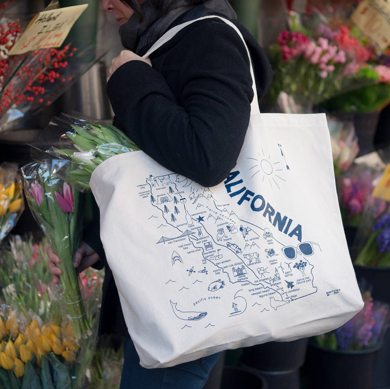 Person holding a tote bag with a map of California design in a flower market.