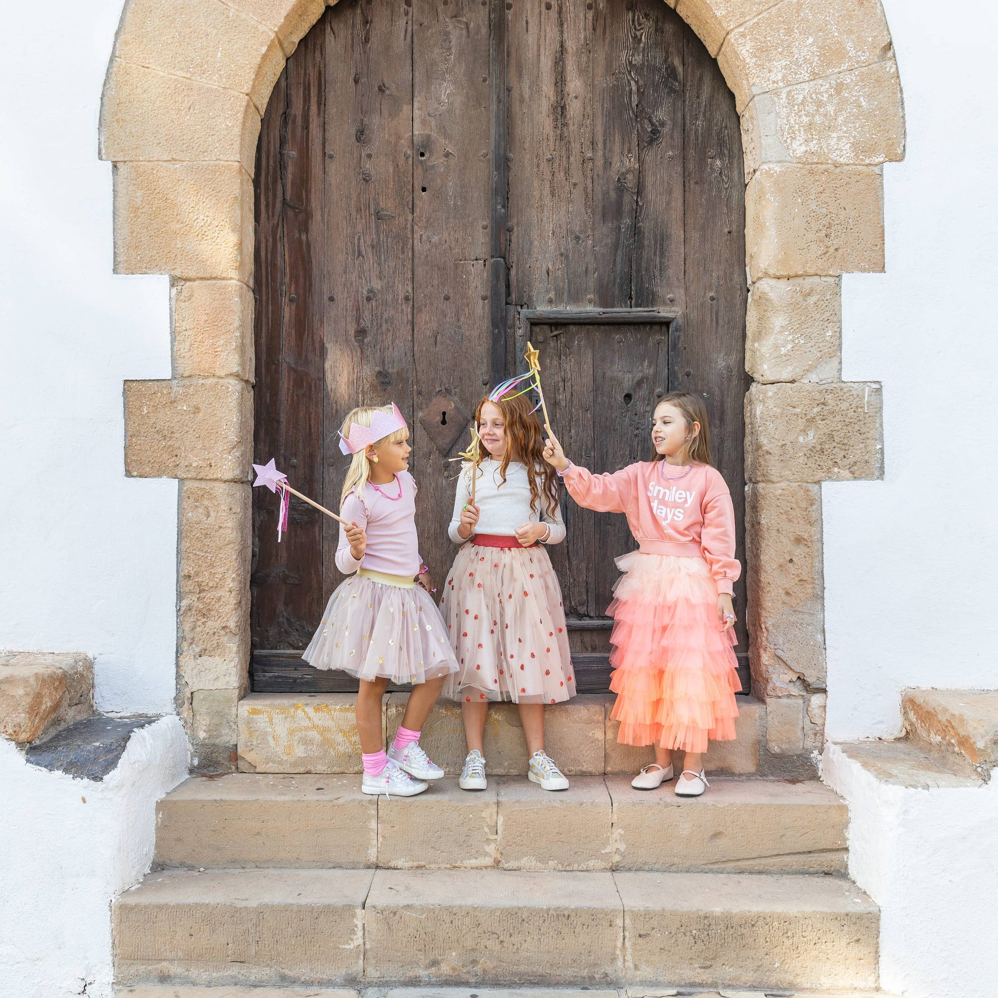 Three young girls in colorful dresses standing in front of a large wooden door with stone archway.