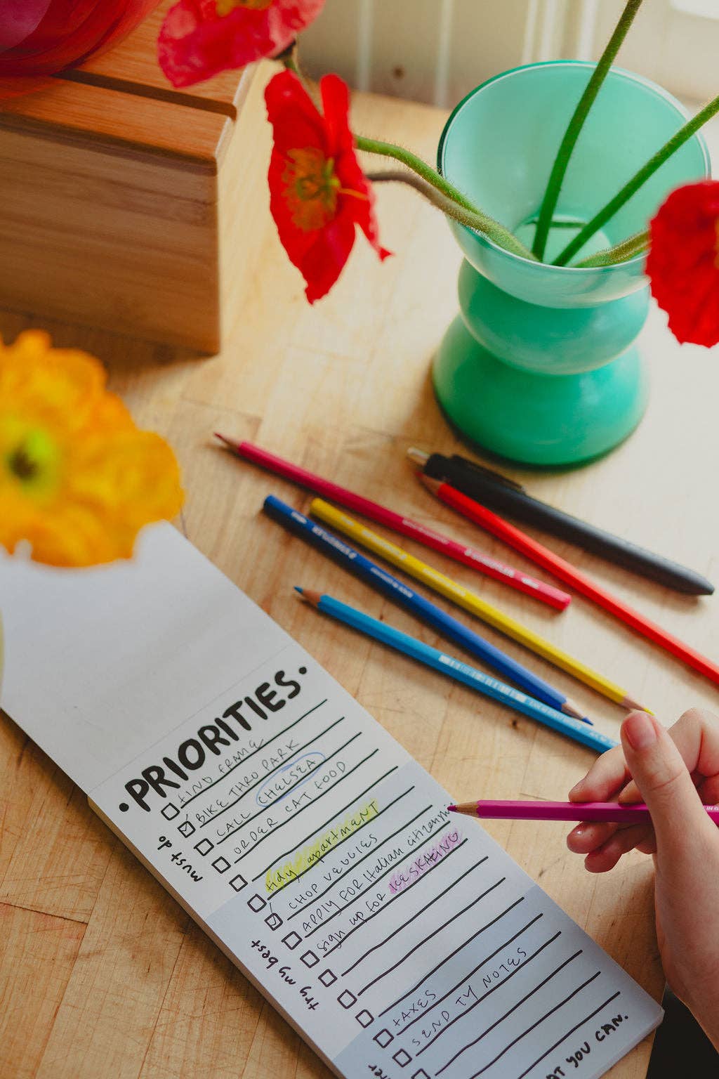 Person filling out a 'Priorities' checklist with colorful pencils on a wooden table with flowers and a green vase.