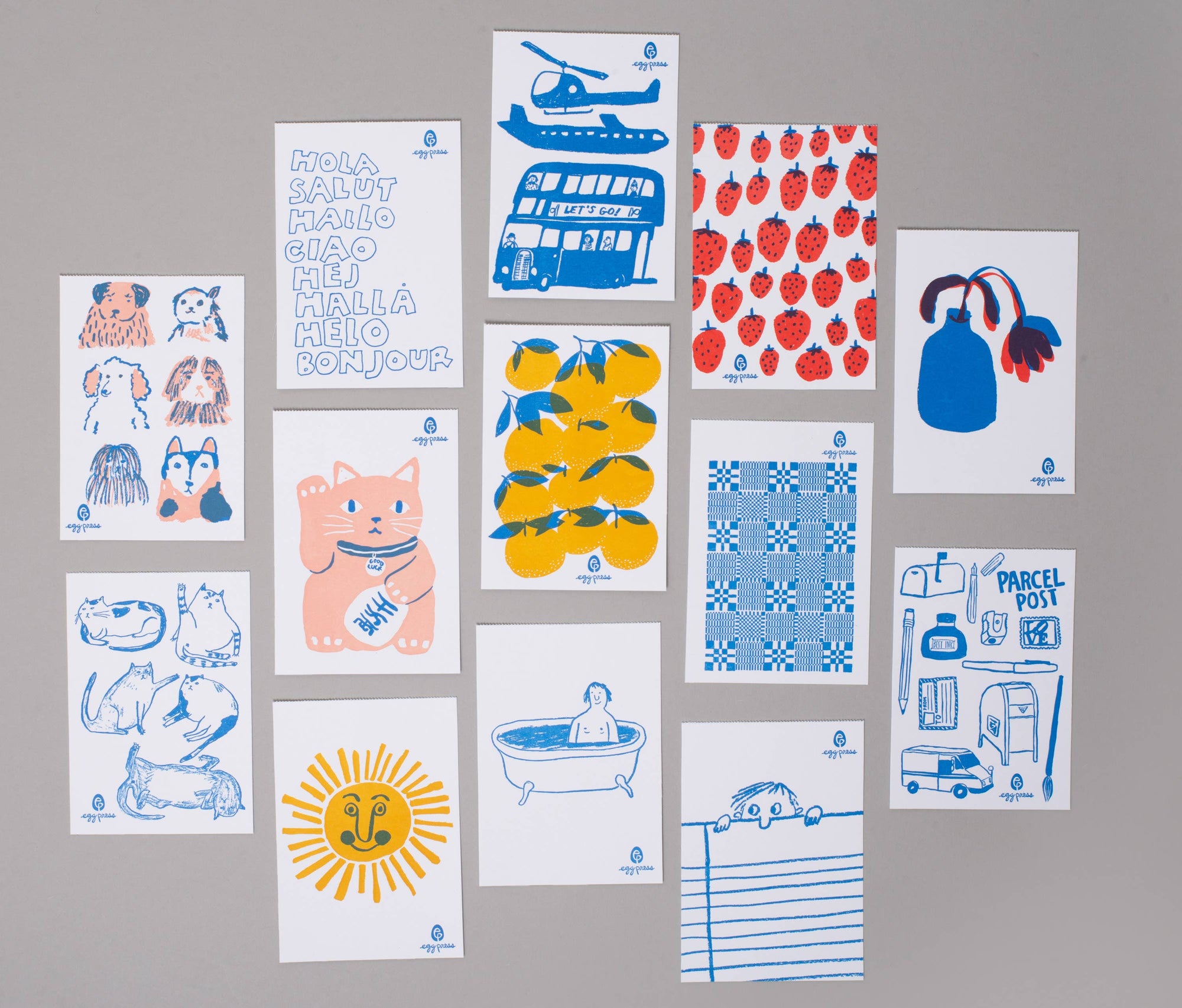 A set of twelve white art prints with blue and red illustrations, arranged in a grid on a neutral gray background. The prints feature whimsical drawings including a helicopter, strawberries, lemons, a bear, a sun, and a parcel box.