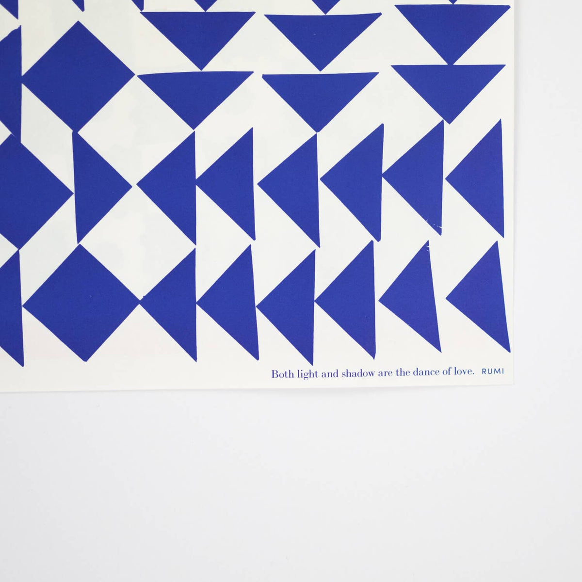A close-up of a white art print featuring a bold geometric pattern of blue triangles and diamonds, with the text 'Both light and shadow are the dance of love, RUMI' printed in blue at the bottom.