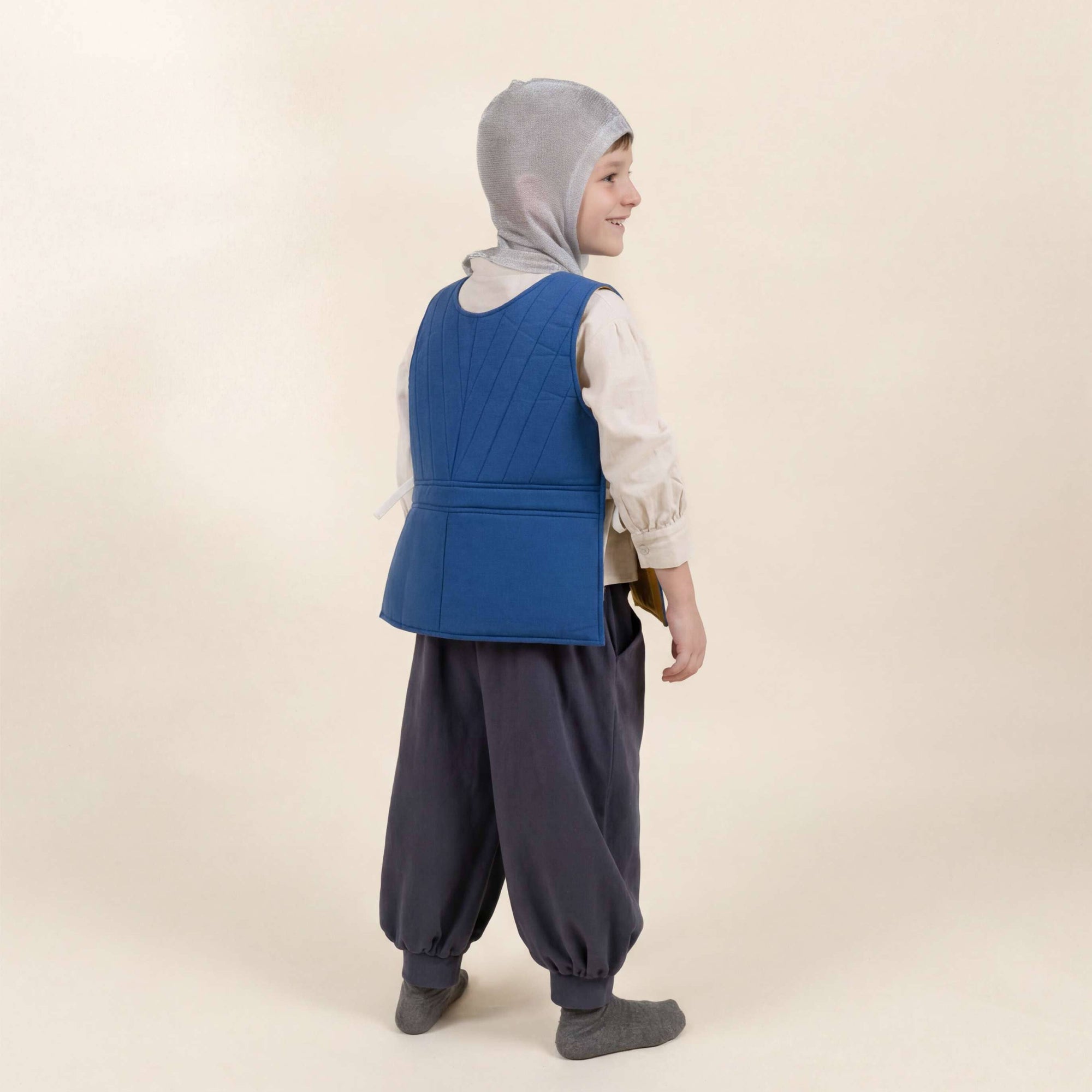 Person wearing a blue vest, white shirt, gray hijab, and dark pants on a beige background