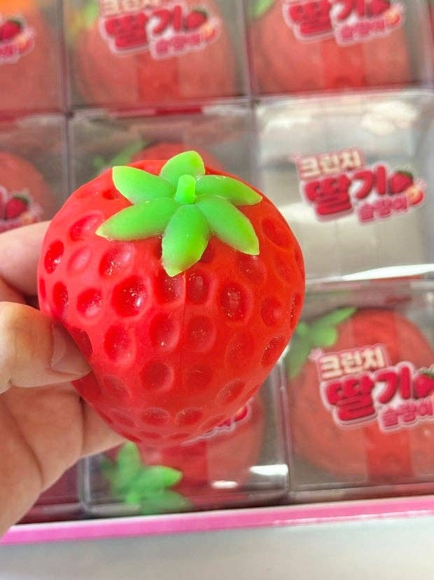 A hand holding a red strawberry-shaped squishy toy with green leaves, with packaged strawberry squishies in the background.