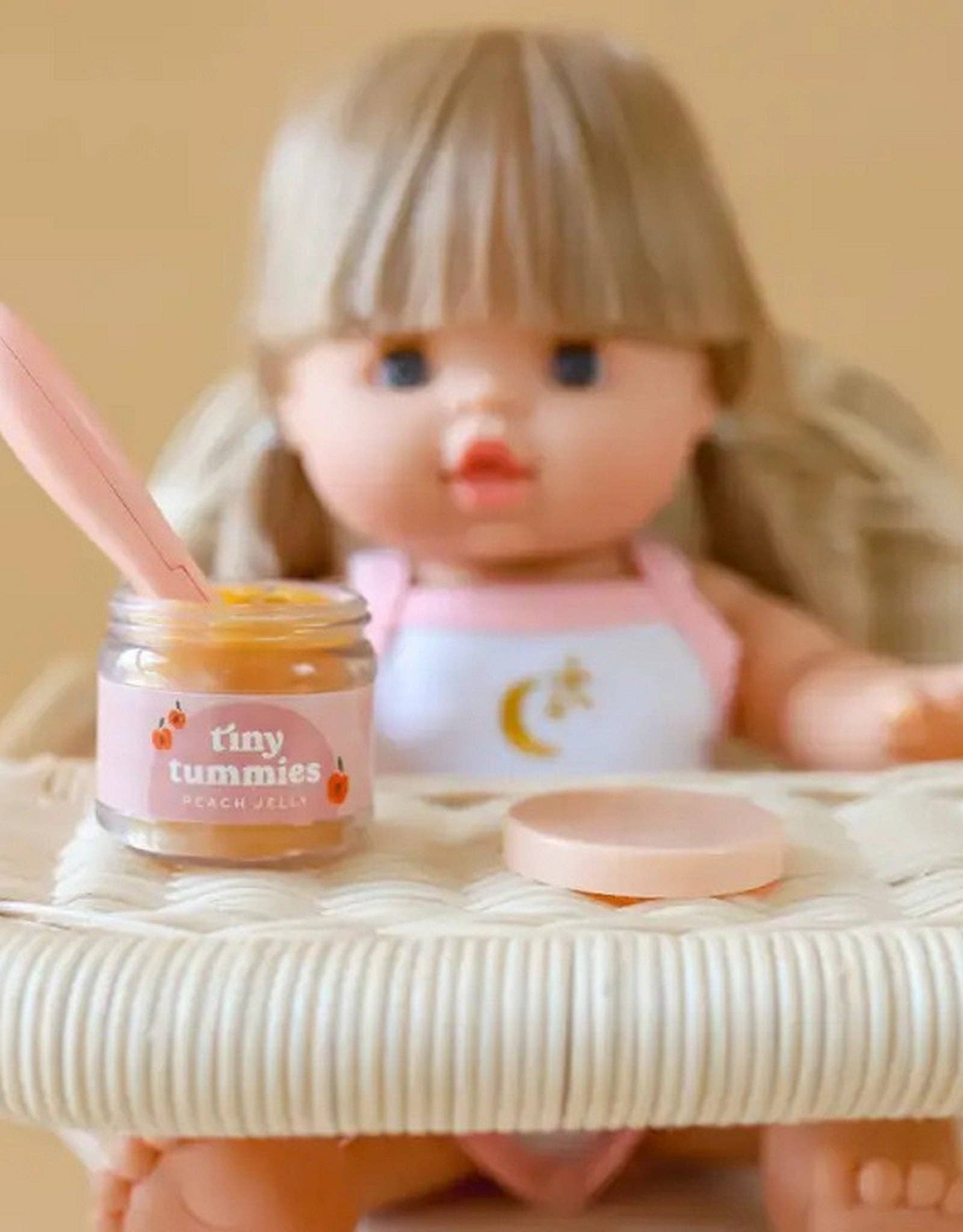 Small jar labeled 'tiny tummies' next to a doll on a white surface