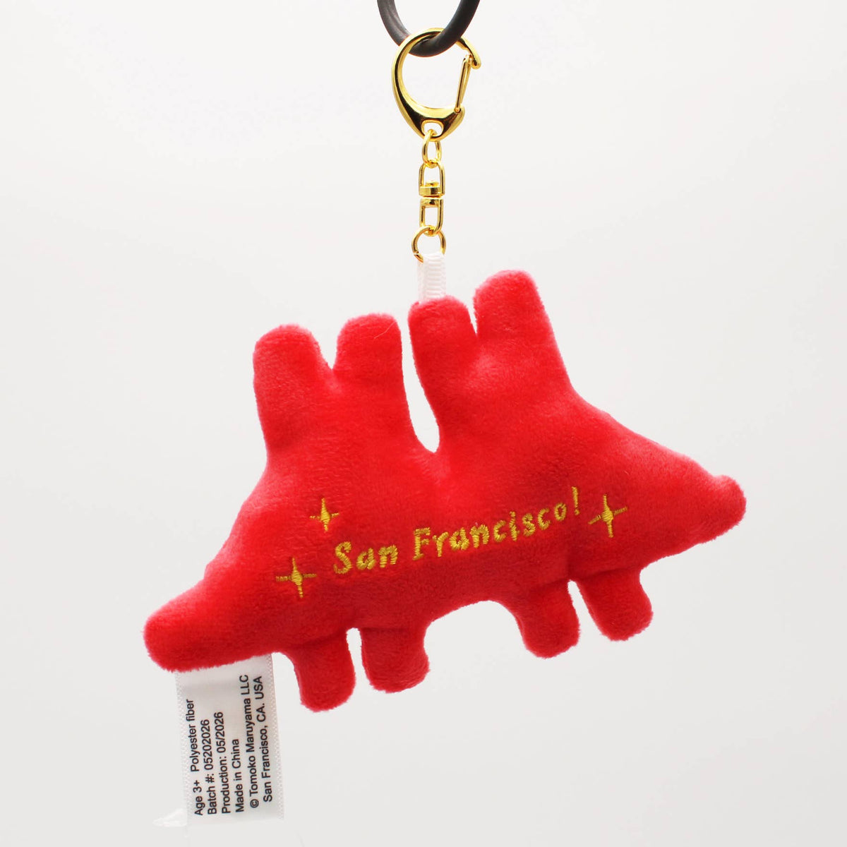 A red plush dinosaur-shaped keychain charm with 'San Francisco!' embroidered in yellow on the side, attached to a gold metal clasp and black strap, shown against a plain white background.
