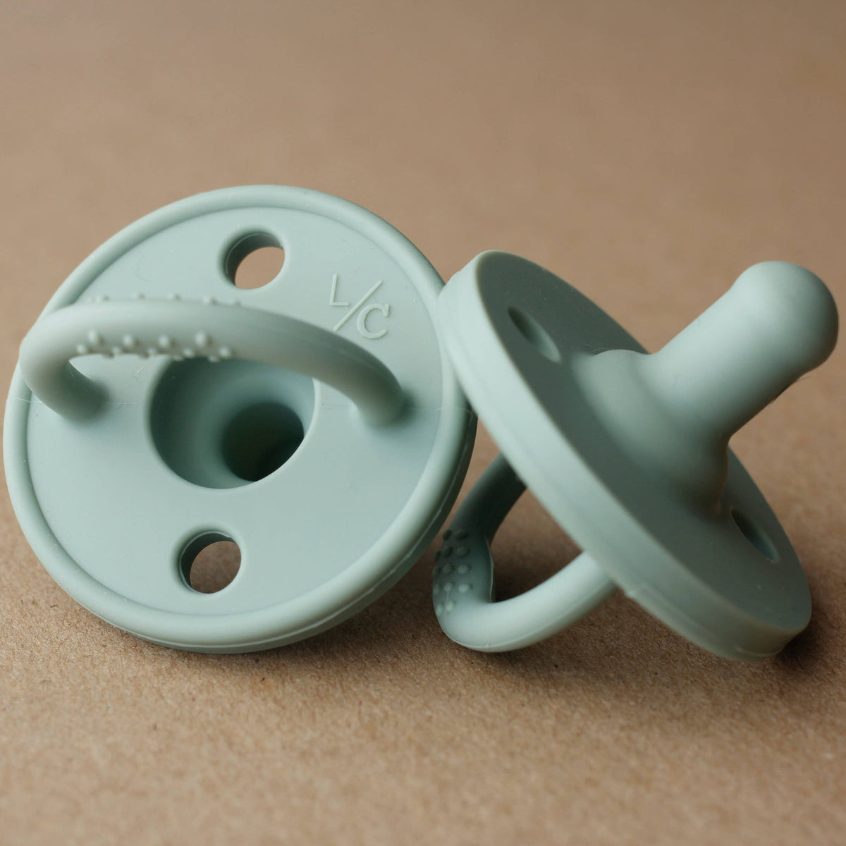 Two sage green silicone baby pacifiers with teething grips, one standing upright and the other lying on its side, set against a textured brown background.