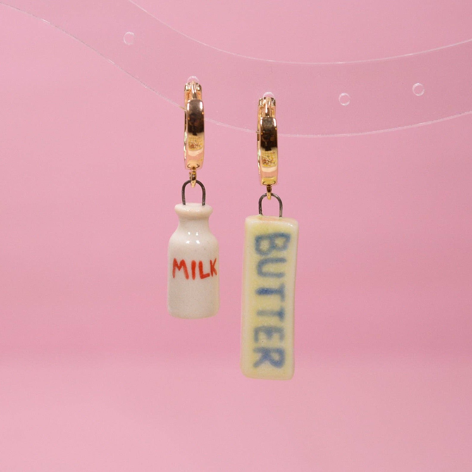 Earrings resembling small bottles labeled 'Milk' and 'Butter' on a pink background