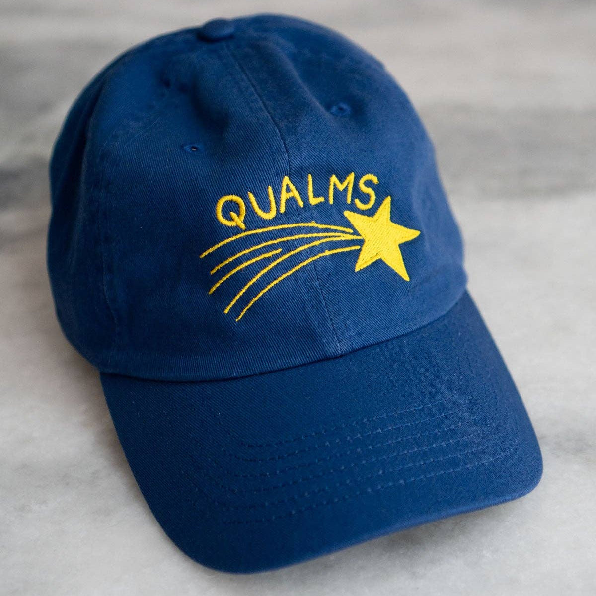 Blue cap with 'QUALMS' and a star design on a gray surface