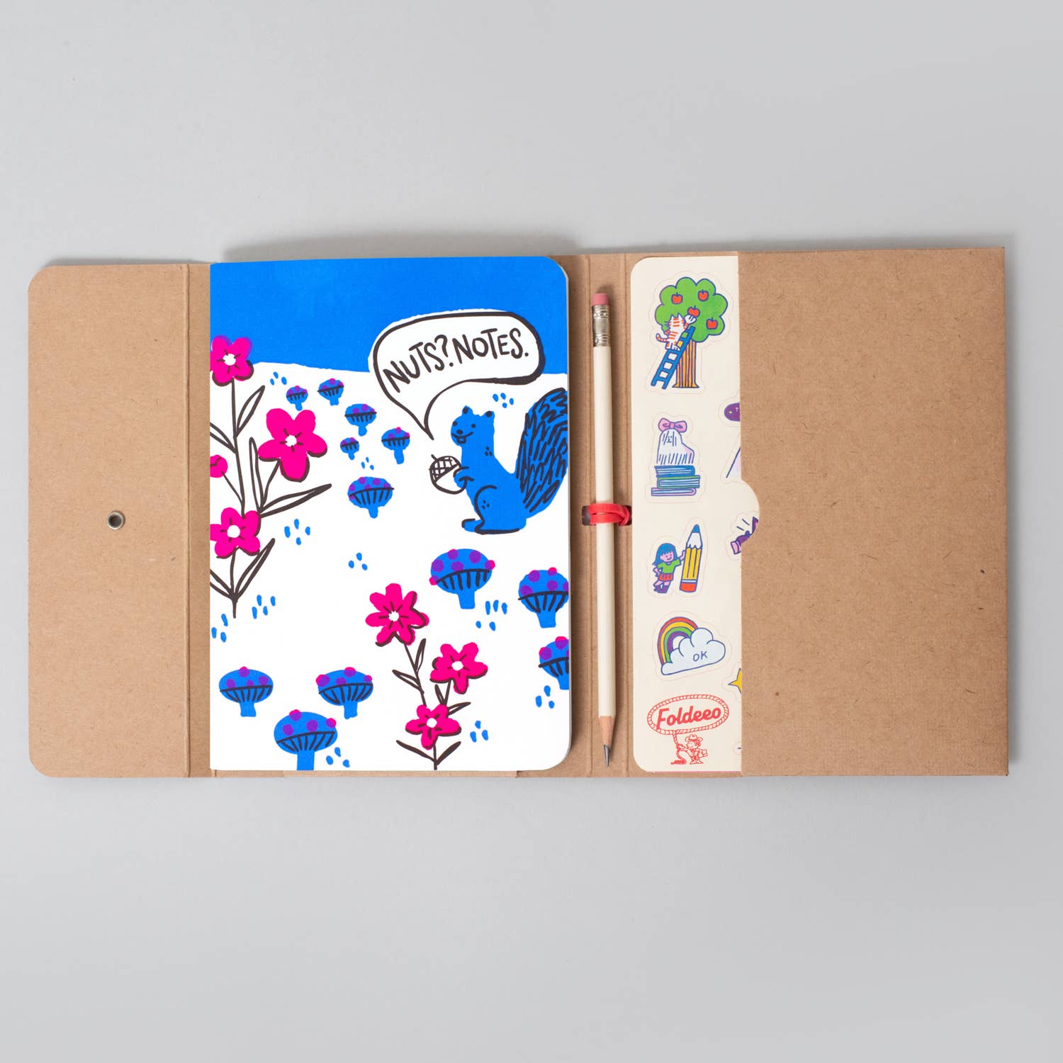 A kraft paper notebook cover opened flat on a light gray background, showing a white notebook insert with a blue squirrel illustration, pink flowers, mushroom graphics, and a speech bubble reading 'NUTS NOTES.' A pencil and a sticker sheet with colorful doodles are tucked into the right pocket.
