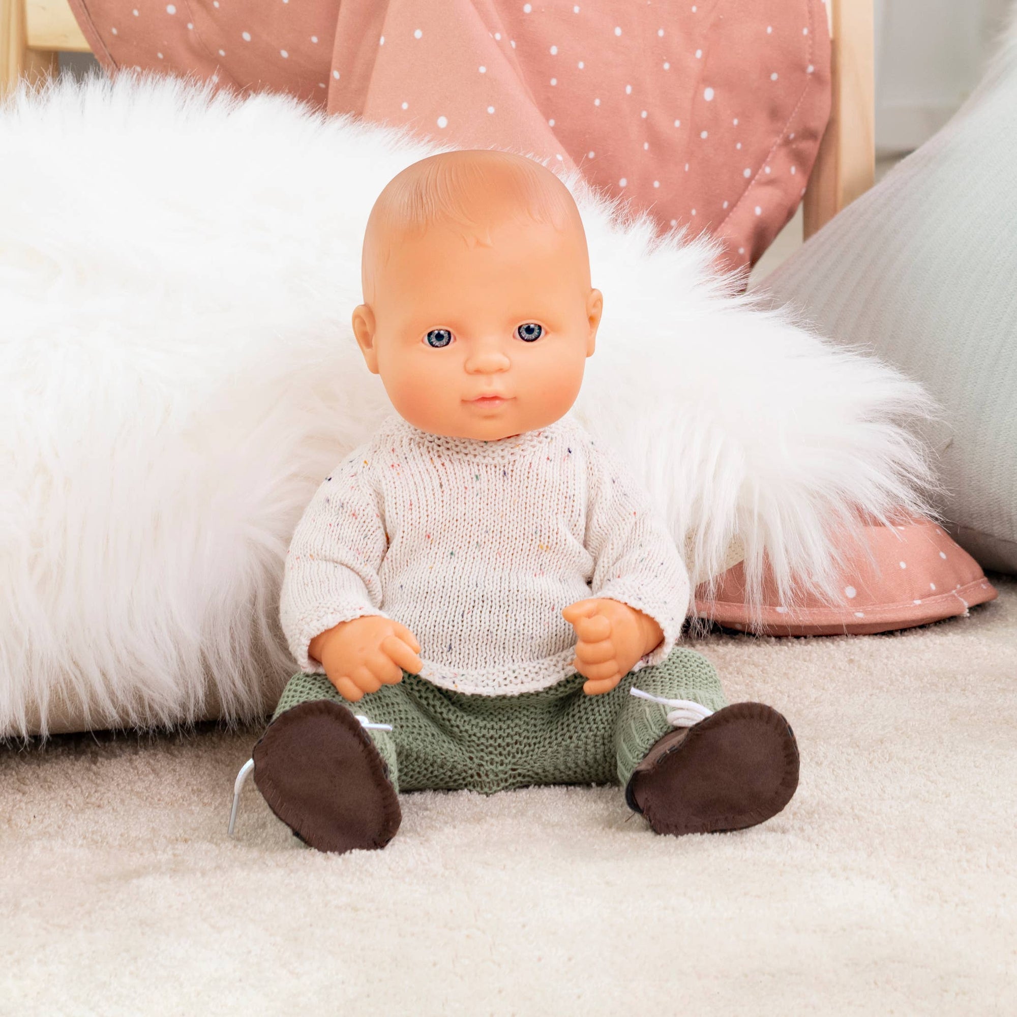 Baby doll wearing a knitted outfit sitting on a soft surface with decorative pillows in the background.