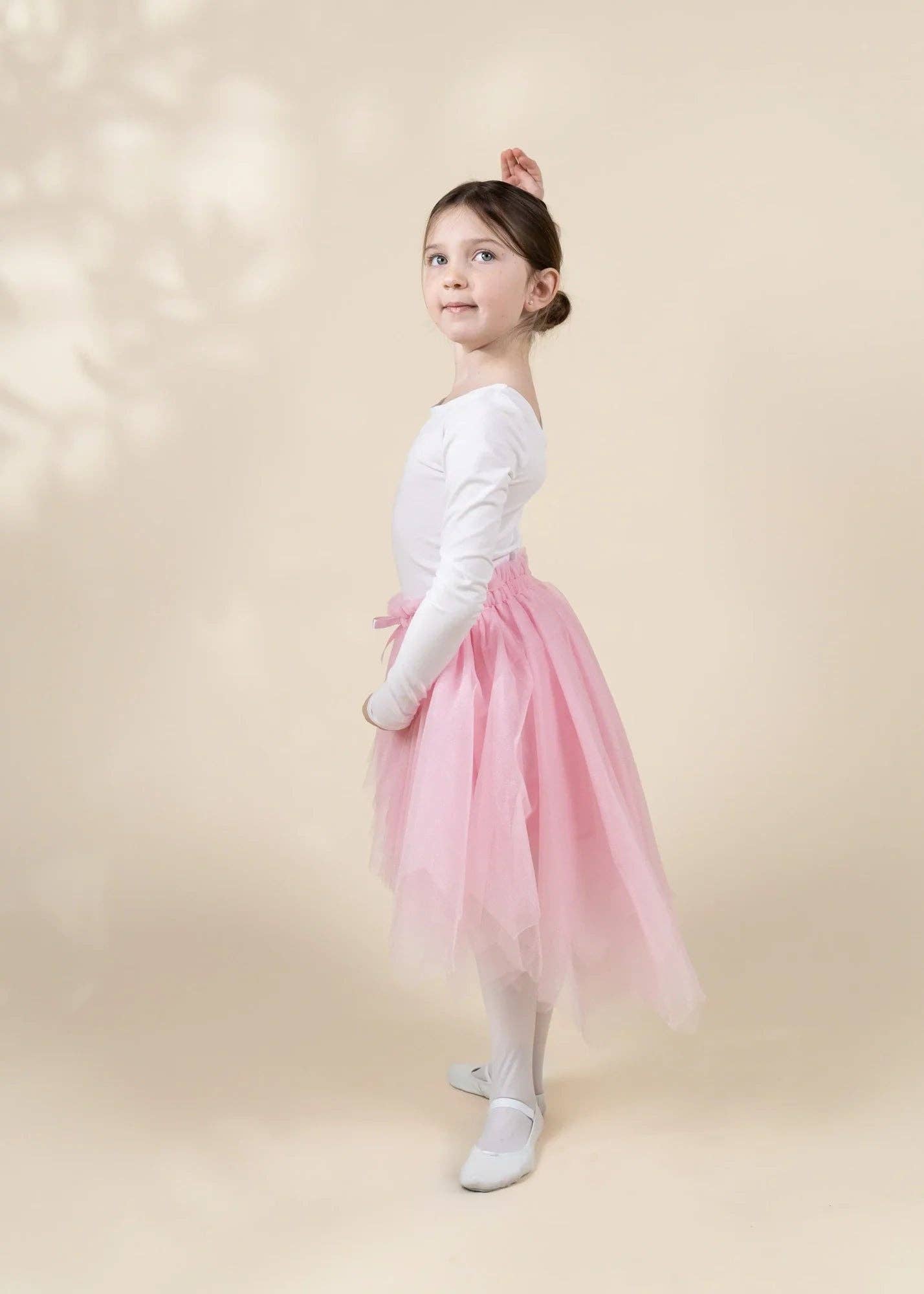 A young girl dancer posing in profile wearing a white long-sleeve leotard, a fluffy pink tutu skirt, white tights, and white ballet shoes against a warm beige background with soft leaf shadows.