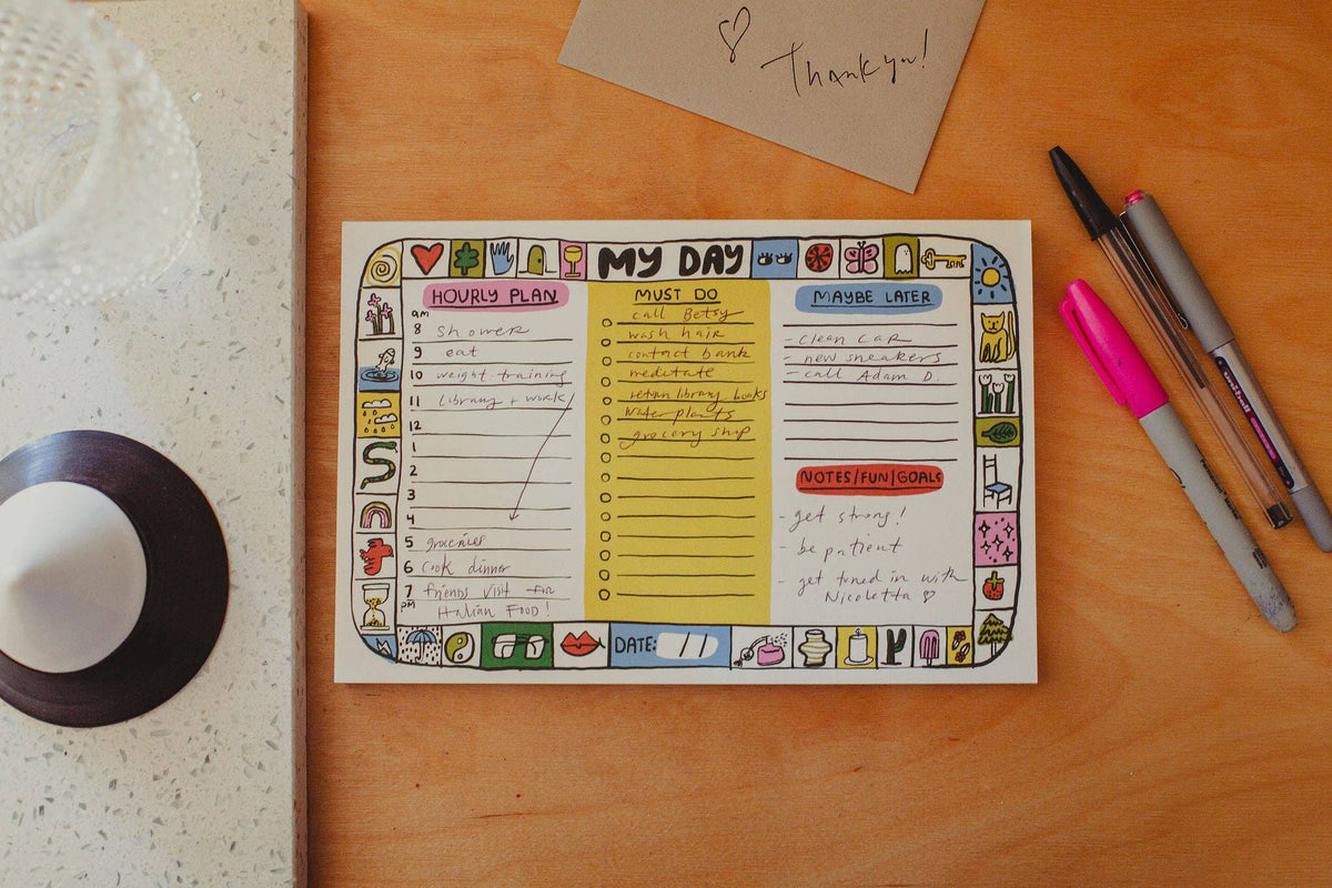 A hand-drawn daily planner page titled 'MY DAY' laid on a wooden tabletop, surrounded by pens, a handwritten thank-you note, and a small decorative object. The planner has colorful illustrated borders and sections for hours plan, must do list, P.