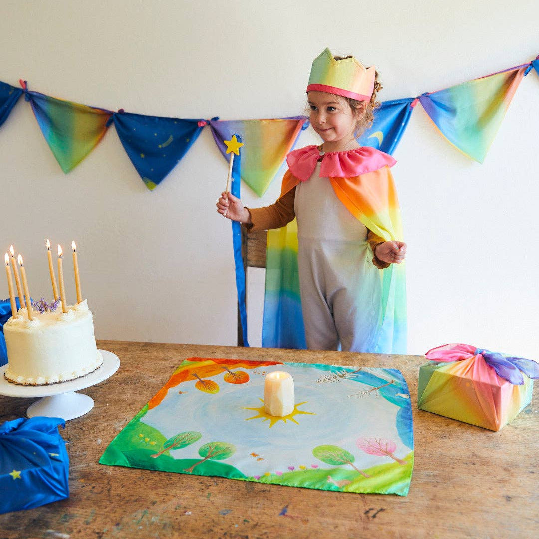 Child in a rainbow-themed costume with a birthday cake and decorations in a room.