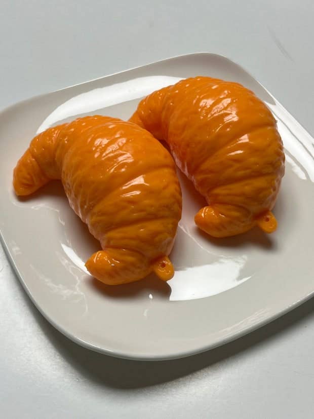 Two glossy orange croissant-shaped items on a white square plate against a light gray background.