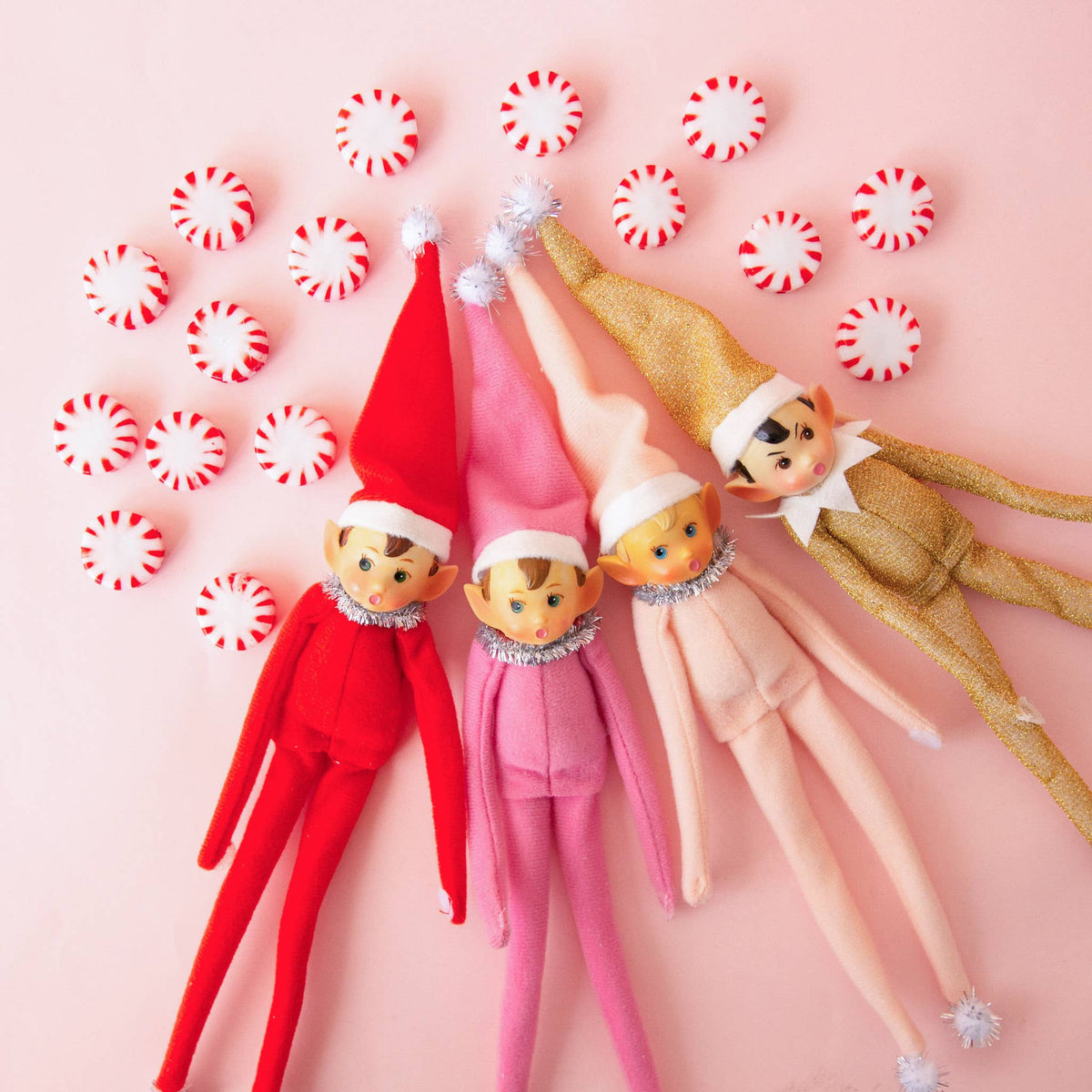 Four plush Elf on a Shelf dolls in red, pink, peach, and gold outfits are arranged on a light pink background with scattered red-and-white peppermint candies.
